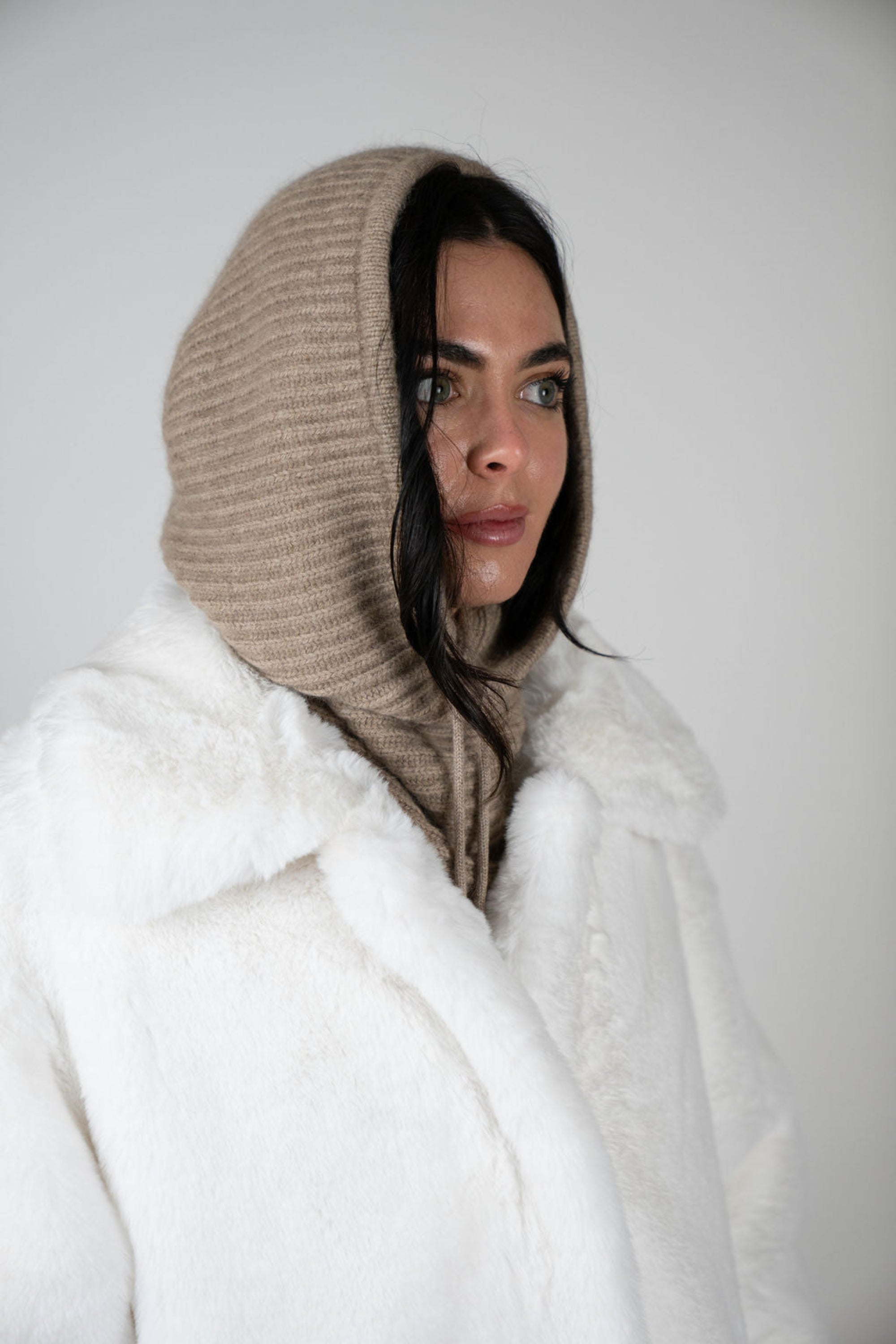 Model wearing the Chalet Knit Hood in Beige by Angel Wings by Lucy, styled with a faux fur vest for elevated winter and luxury resort layering.