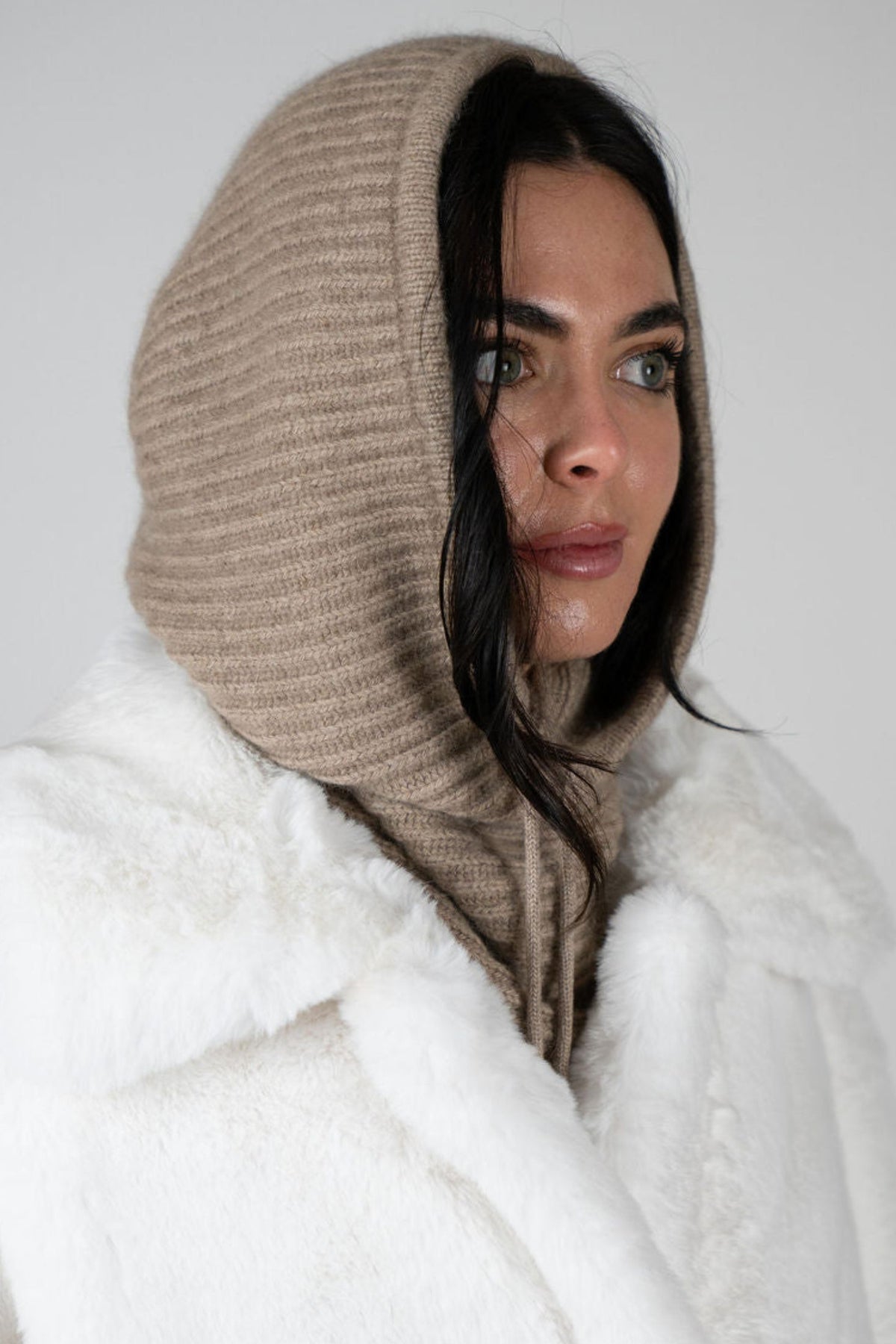 Model wearing the Chalet Knit Hood in Beige by Angel Wings by Lucy, styled with a faux fur vest for elevated winter and luxury resort layering.