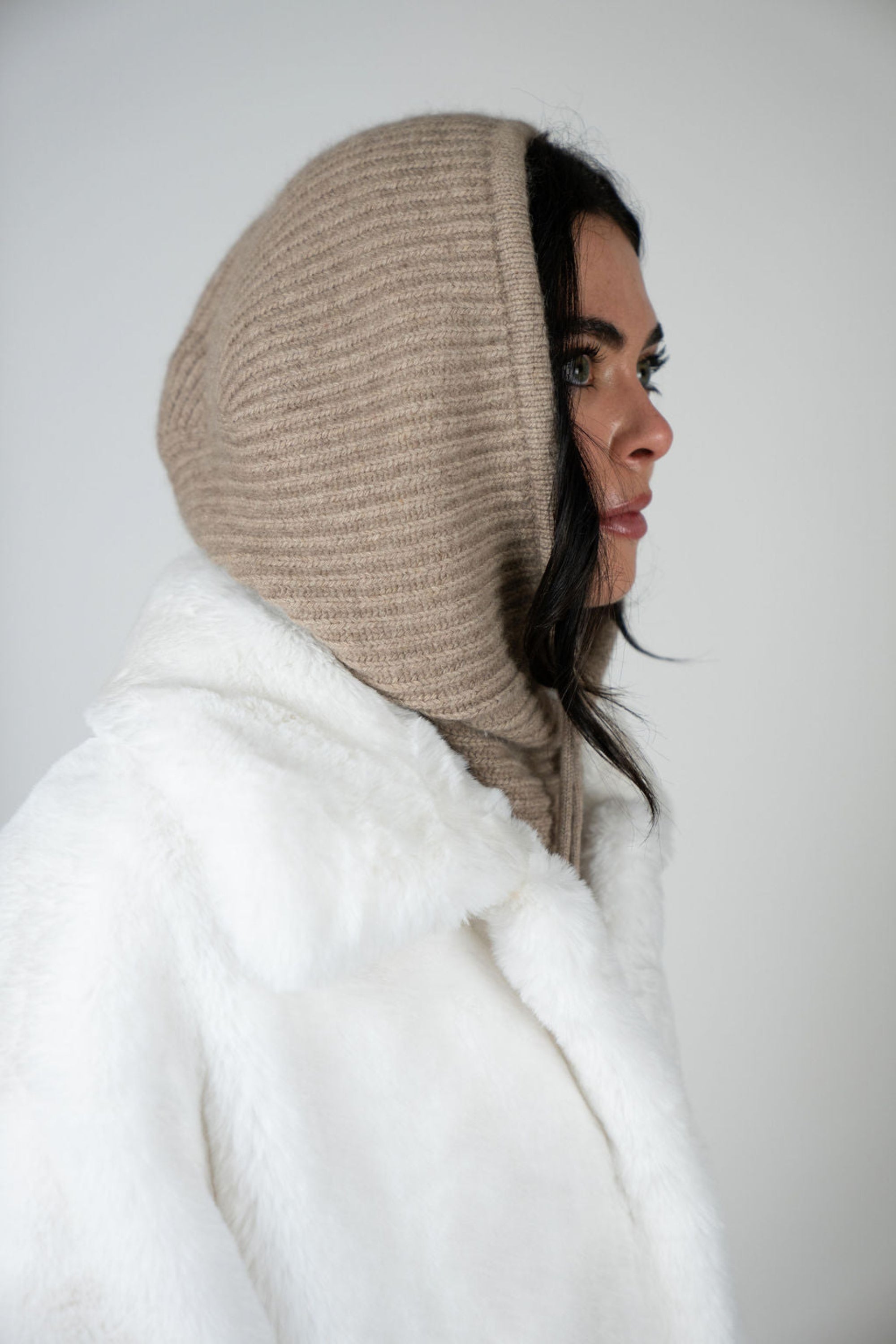 Model wearing the Chalet Knit Hood in Beige by Angel Wings by Lucy, styled with a faux fur vest for elevated winter and luxury resort layering.