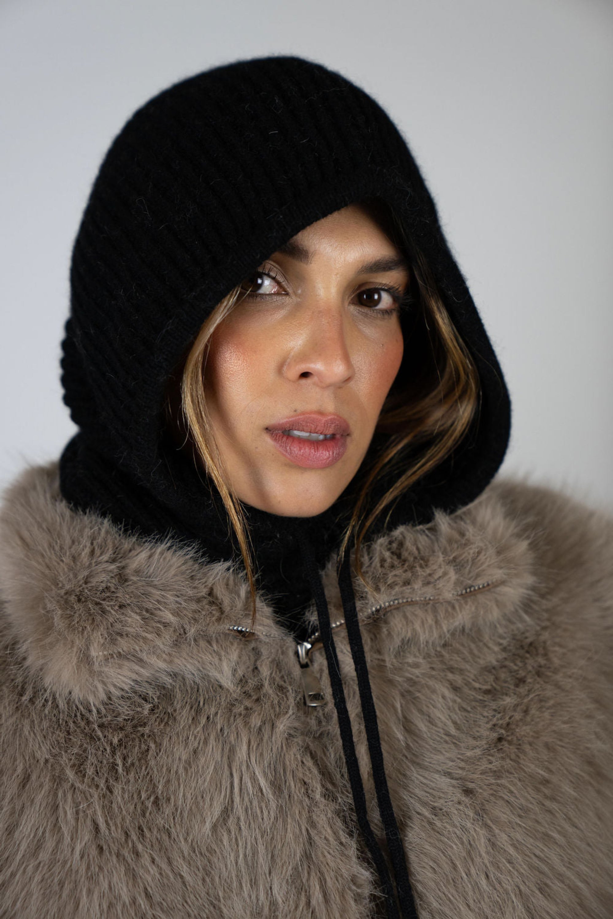 Model wearing the Chalet Knit Hood in Black by Angel Wings by Lucy, styled with a faux fur vest for refined winter and après-ski layering.