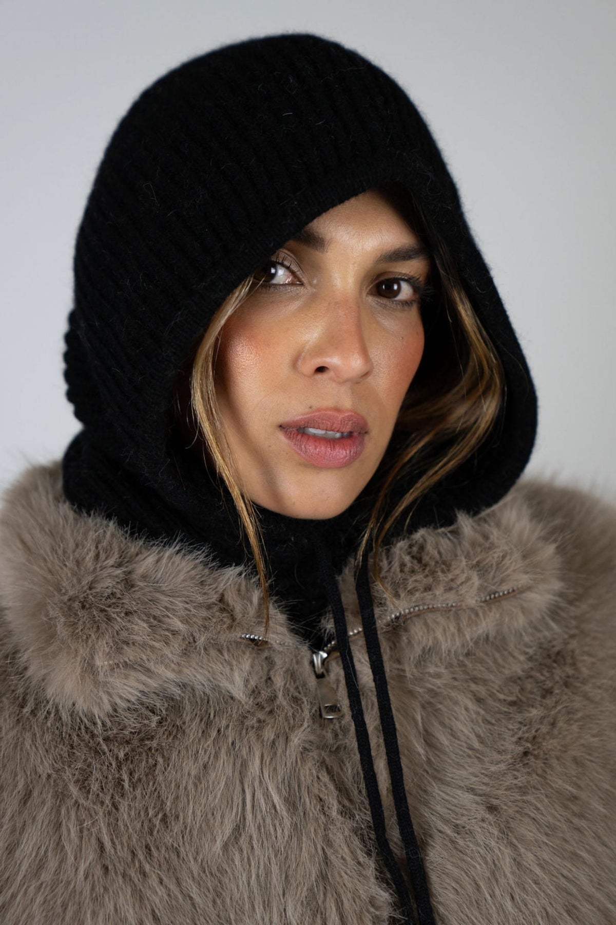 Model wearing the Chalet Knit Hood in Black by Angel Wings by Lucy, styled with a faux fur vest for refined winter and après-ski layering.